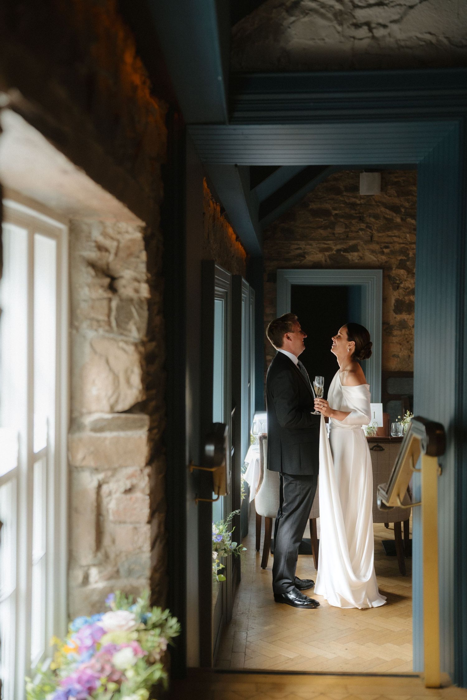 A bride in a white wedding dress stands near a window in a rustic stone-walled room with blue columns.