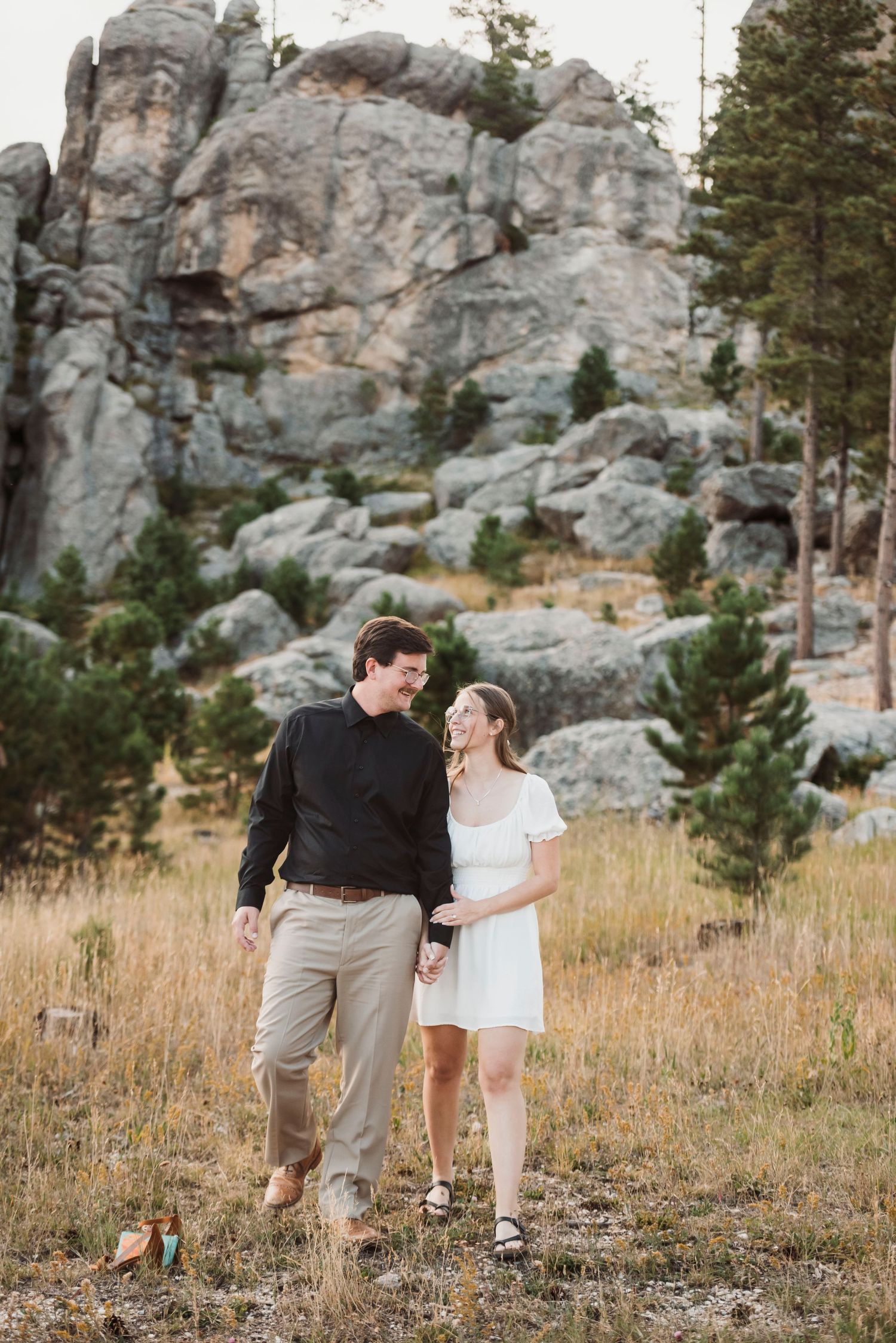 Couple walking together on a grassy hillside with dramatic rocky cliffs in the background.