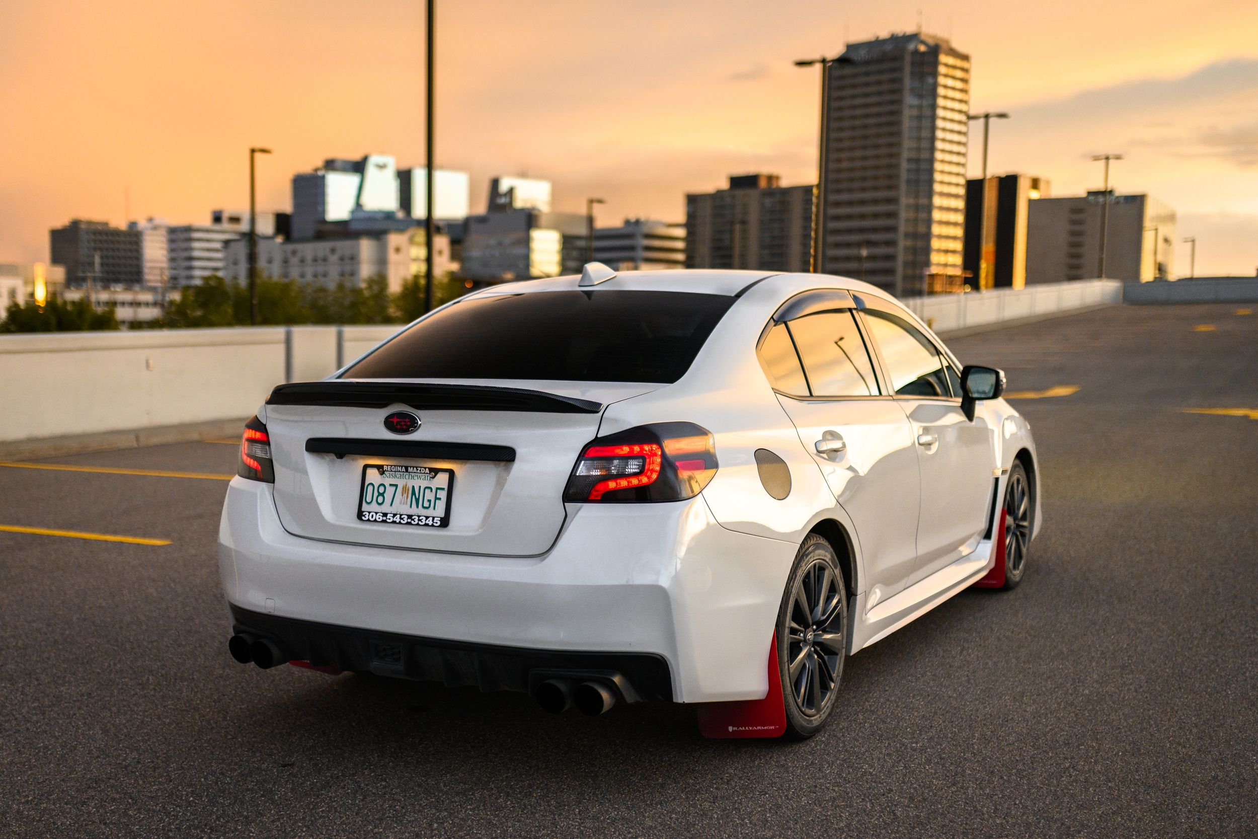 White Subaru WRX STI sedan parked in urban parking lot during sunset with city skyline in background.