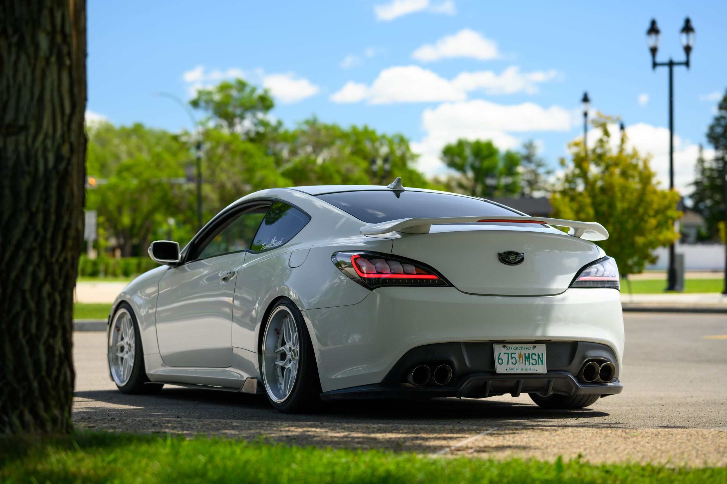 Lowered white Genesis coupe with aftermarket wheels parked near trees.