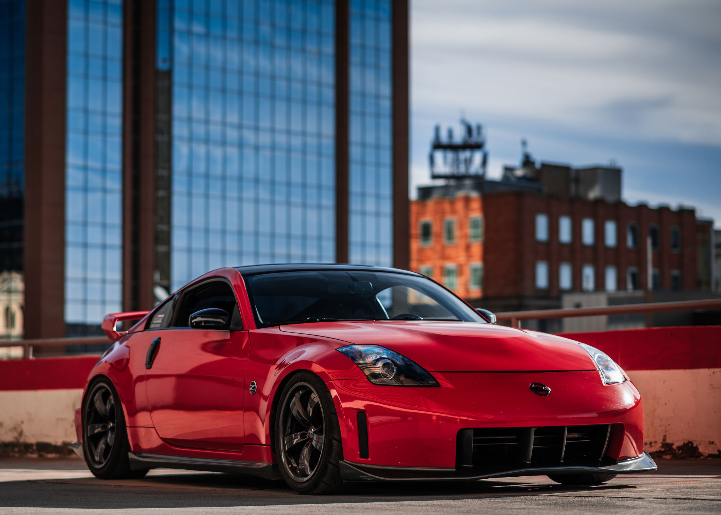 Red Nissan 370Z with aftermarket body kit and wheels against modern building backdrop.