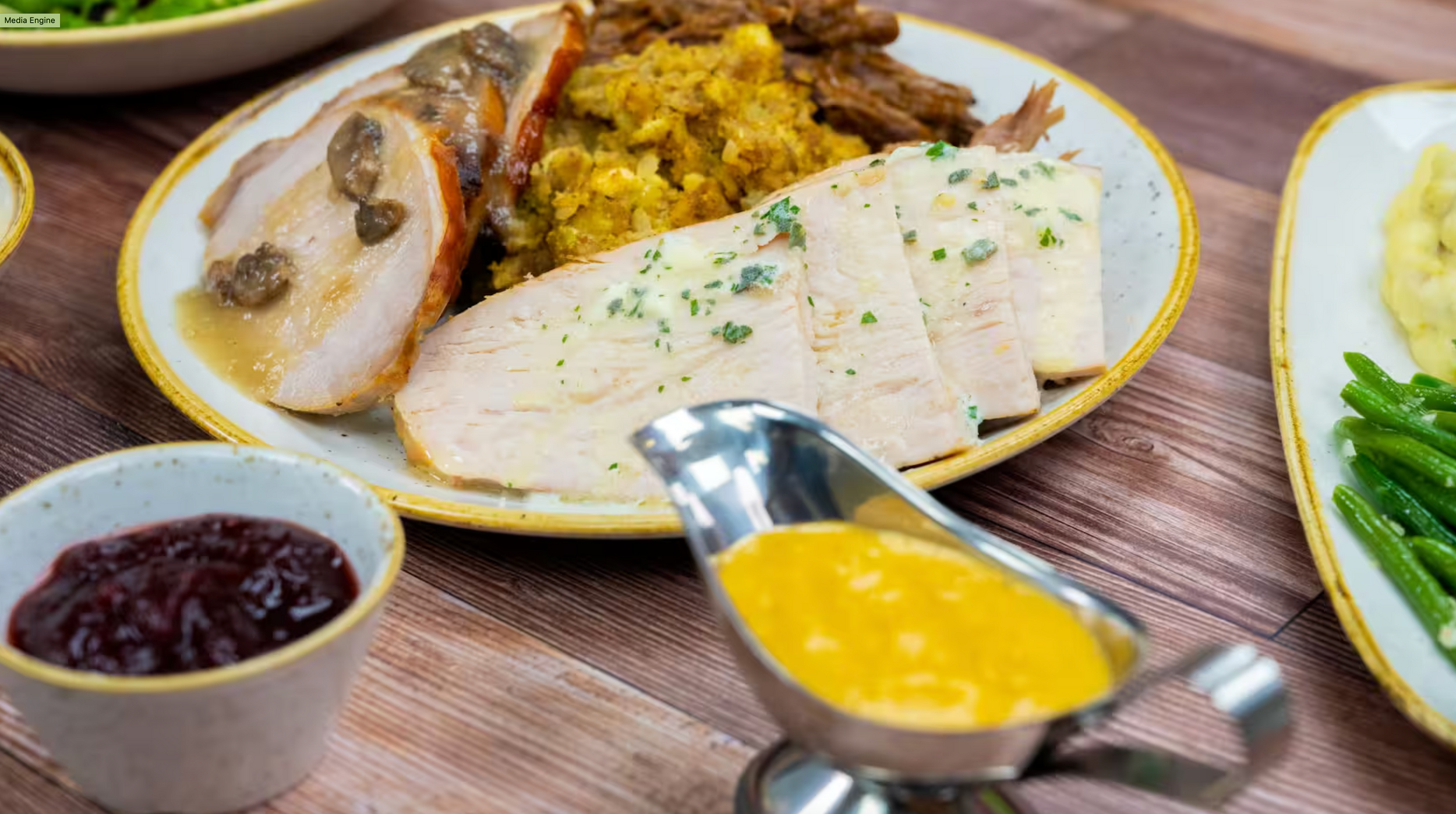 Sliced turkey breast with cranberry sauce, mustard gravy, and stuffing served on a rustic wooden table.