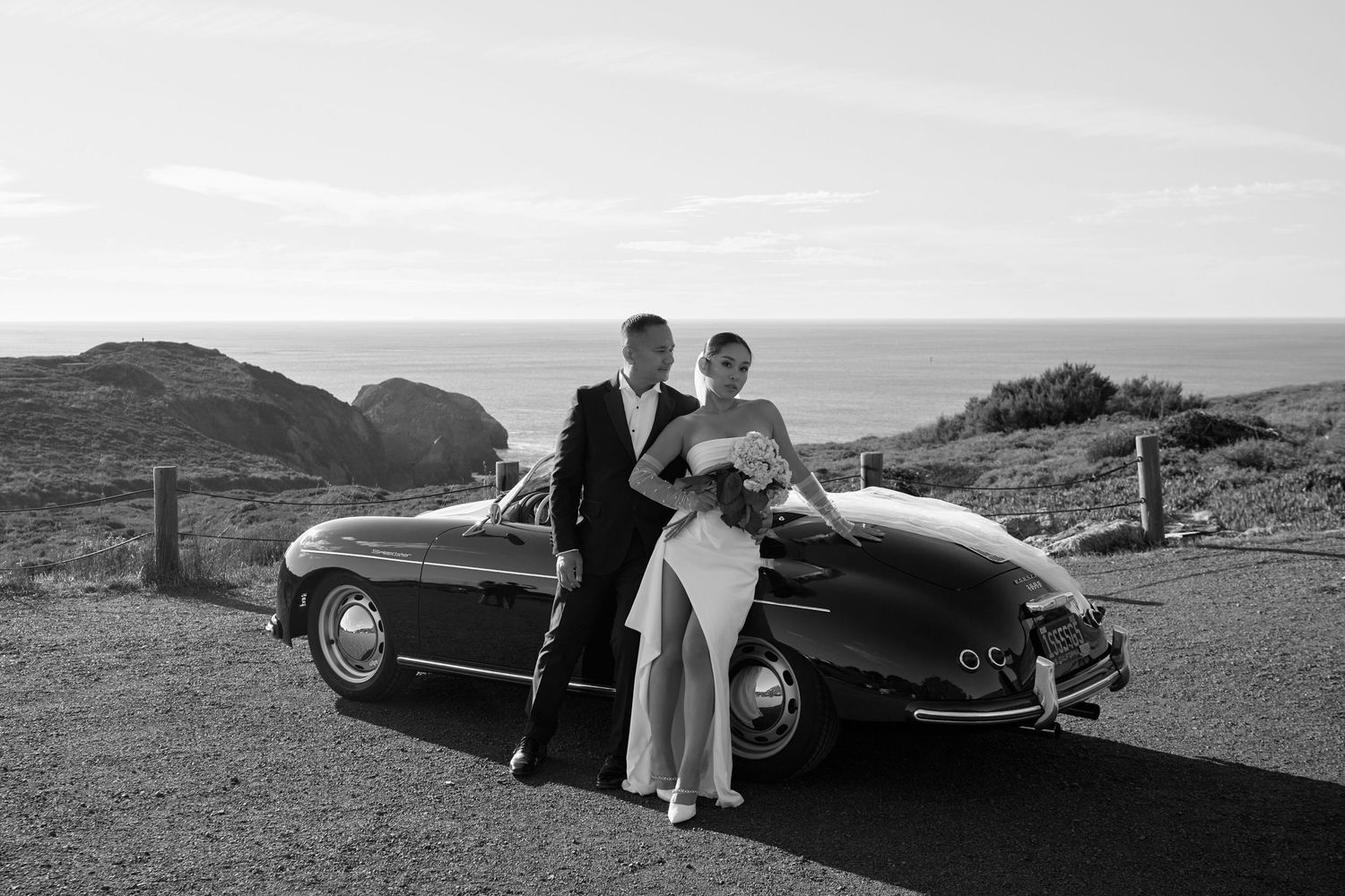 A couple poses with a classic vintage Mercedes convertible on a coastal hilltop overlooking the ocean in black and white.