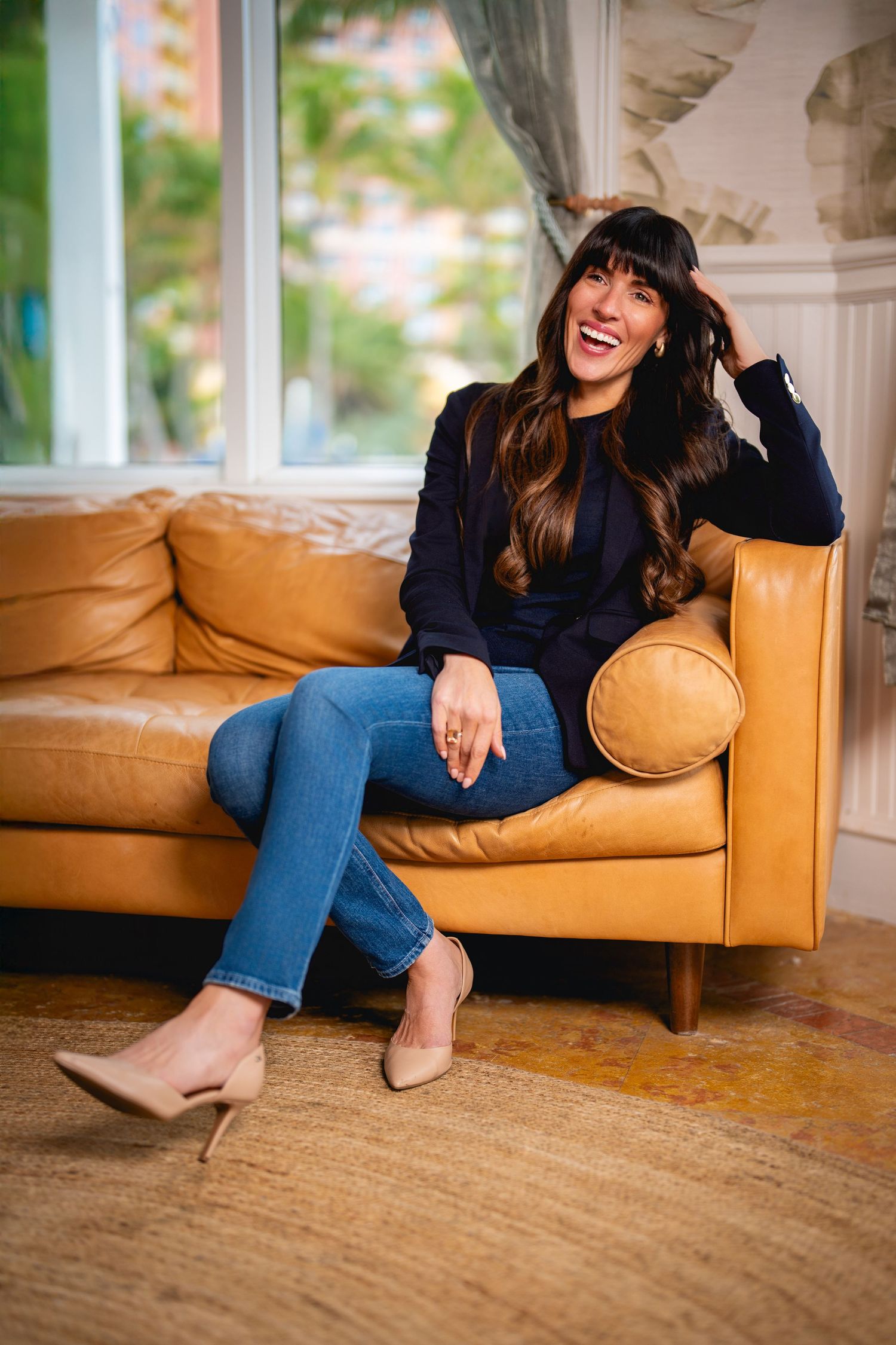 Someone wearing a navy sweater and jeans laughs while sitting casually on a tan leather couch by a bright window.