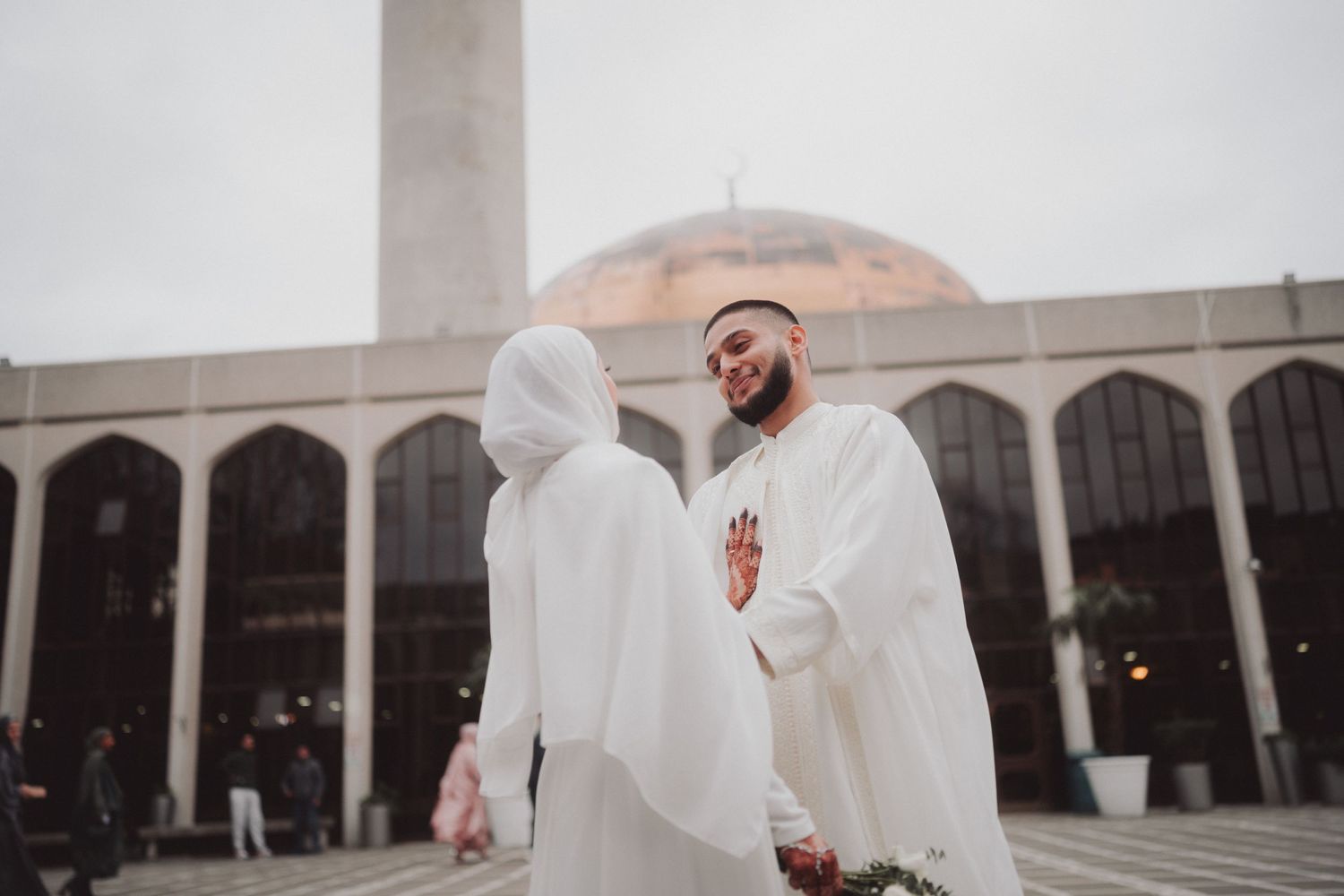 Two people in white traditional dress outside a mosque with domed architecture.