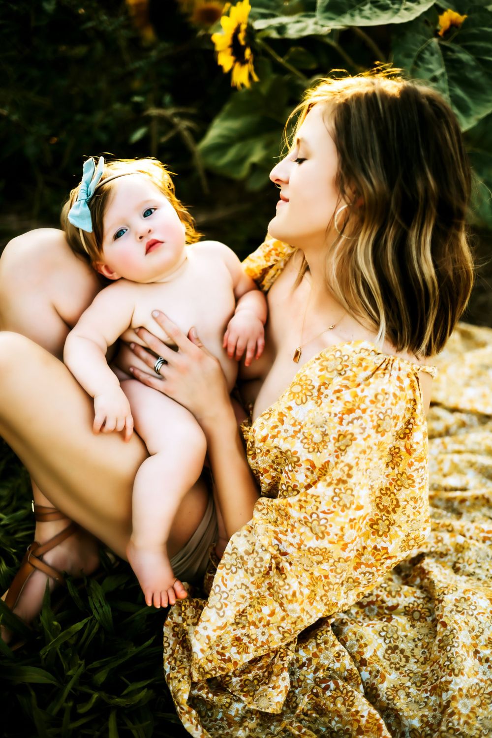 A mother and baby sit together near sunflowers in a garden, with the mother wearing a yellow floral dress.