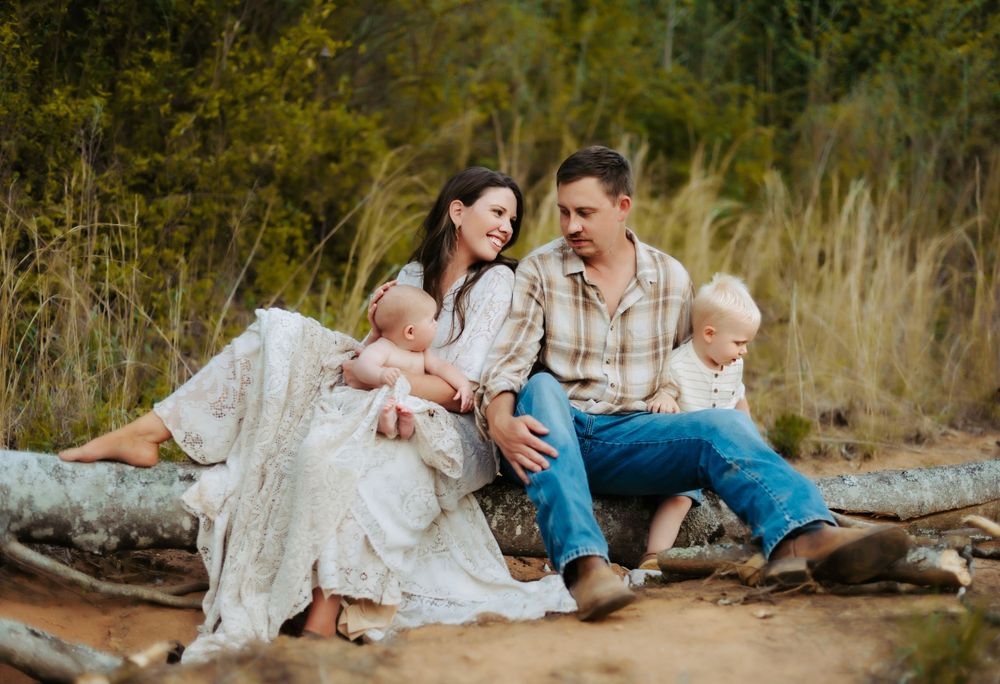 Family in casual attire sits together on a log outdoors with tall grass and natural scenery in background.