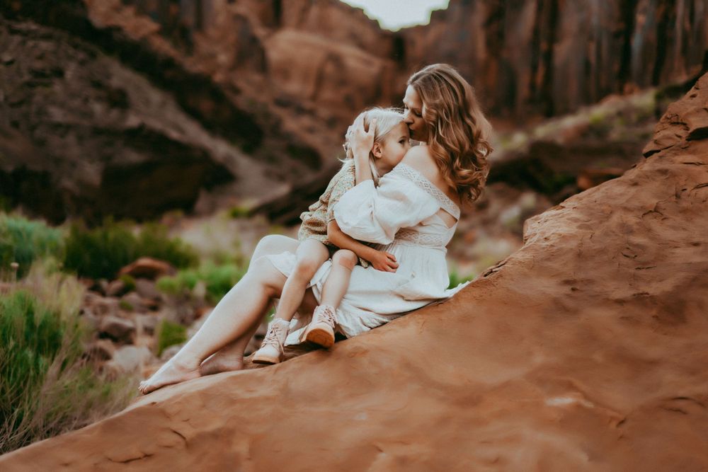 Two people in white outfits sit together on red rock formations at sunset in a desert canyon landscape.