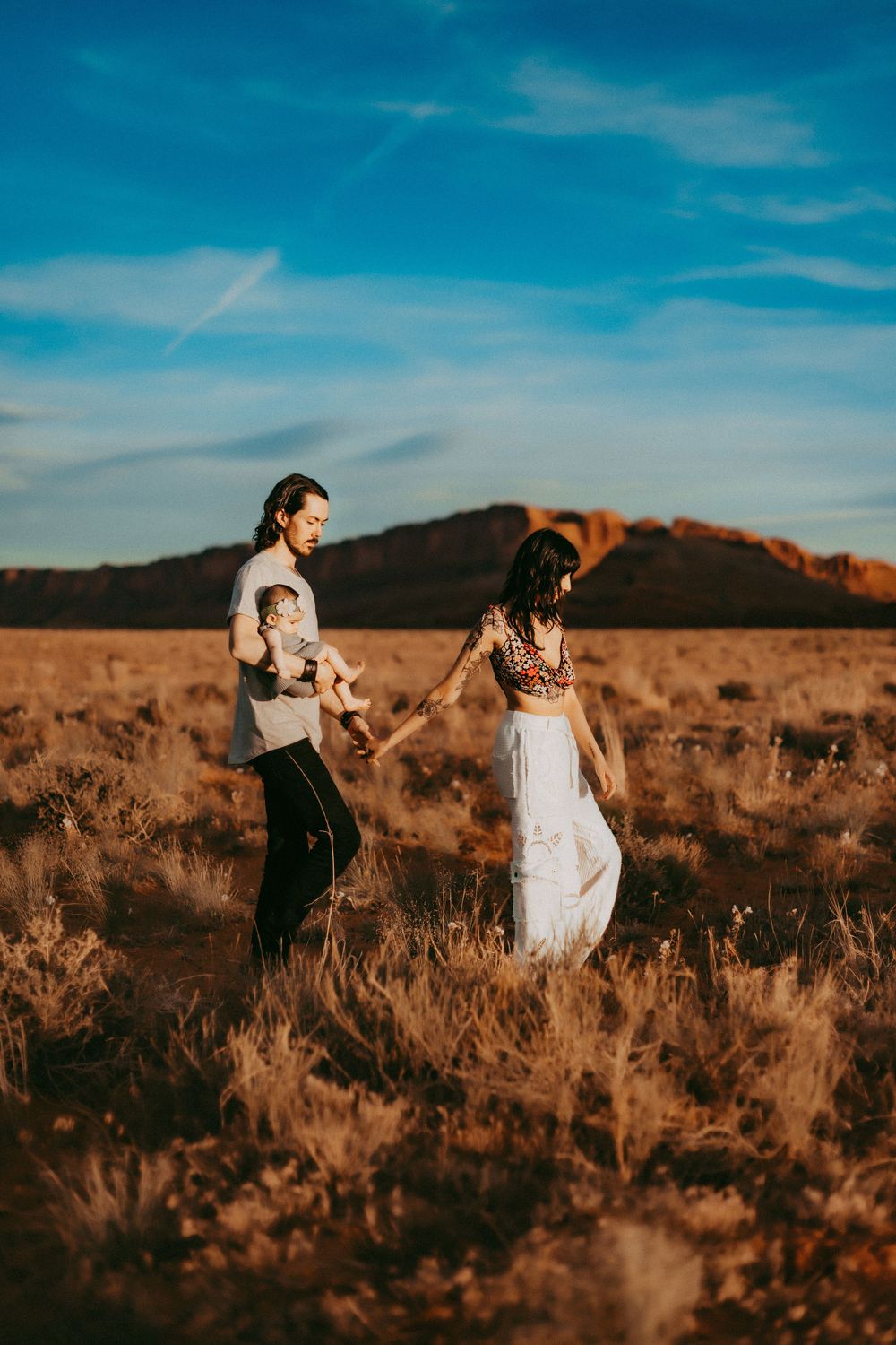 A couple walks hand in hand through a golden desert field at sunset with mountains in the background.