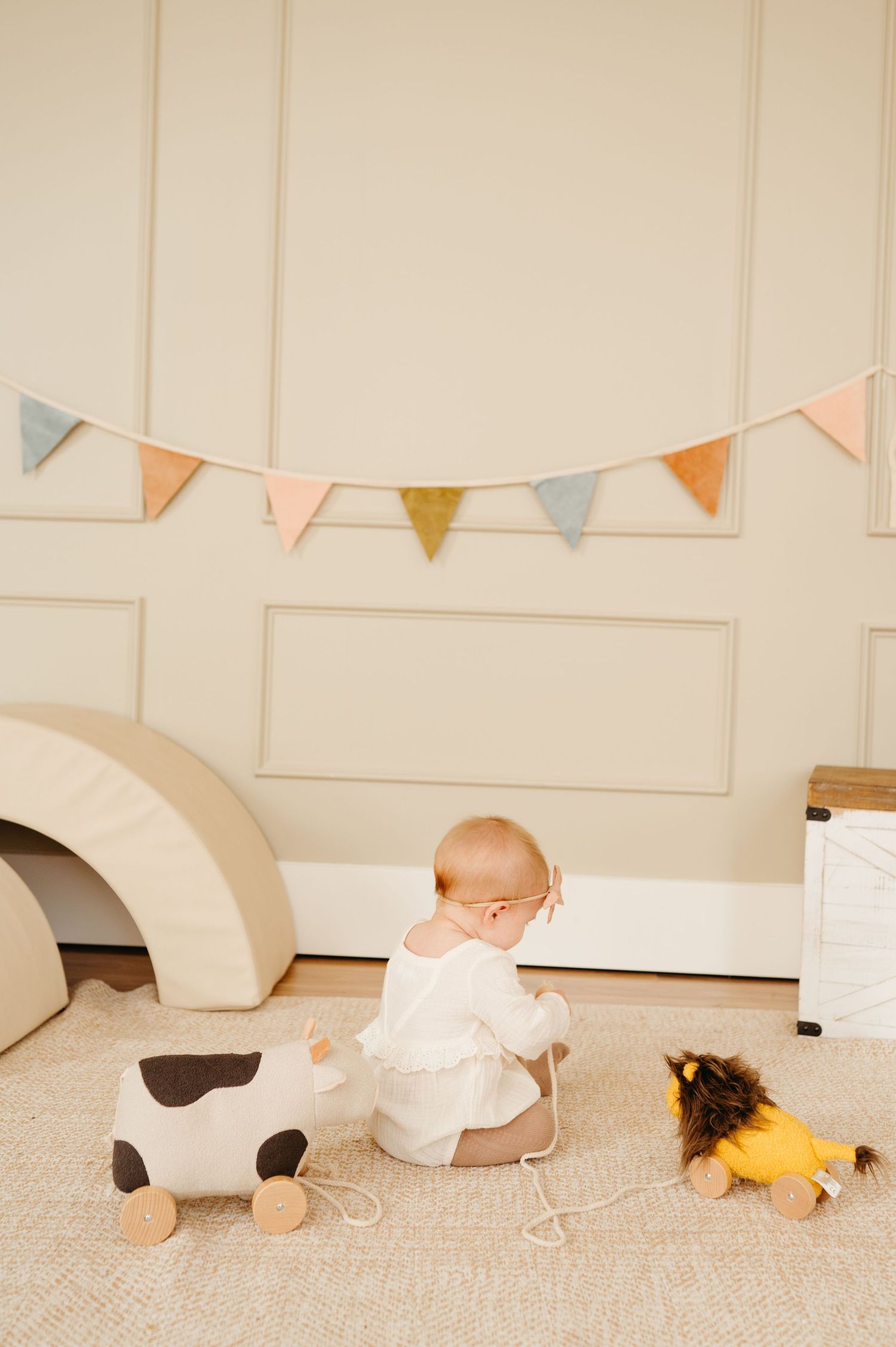 A baby in white sits on a carpet with wooden toys and bunting overhead in a minimalist nursery setting.
