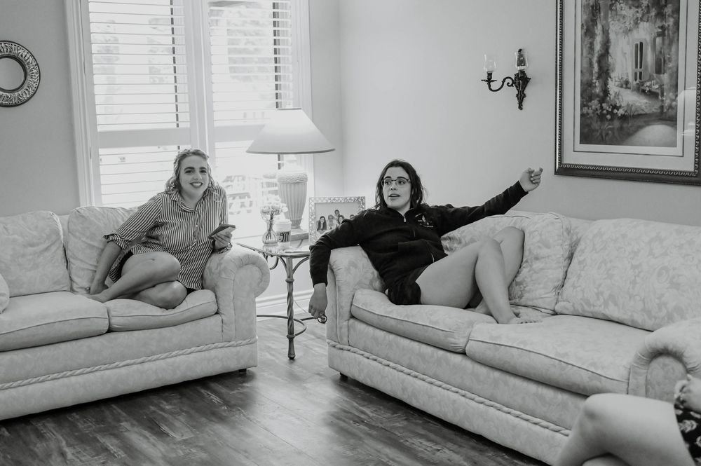 Two people relax casually on separate matching floral sofas in a living room with hardwood floors and white walls.