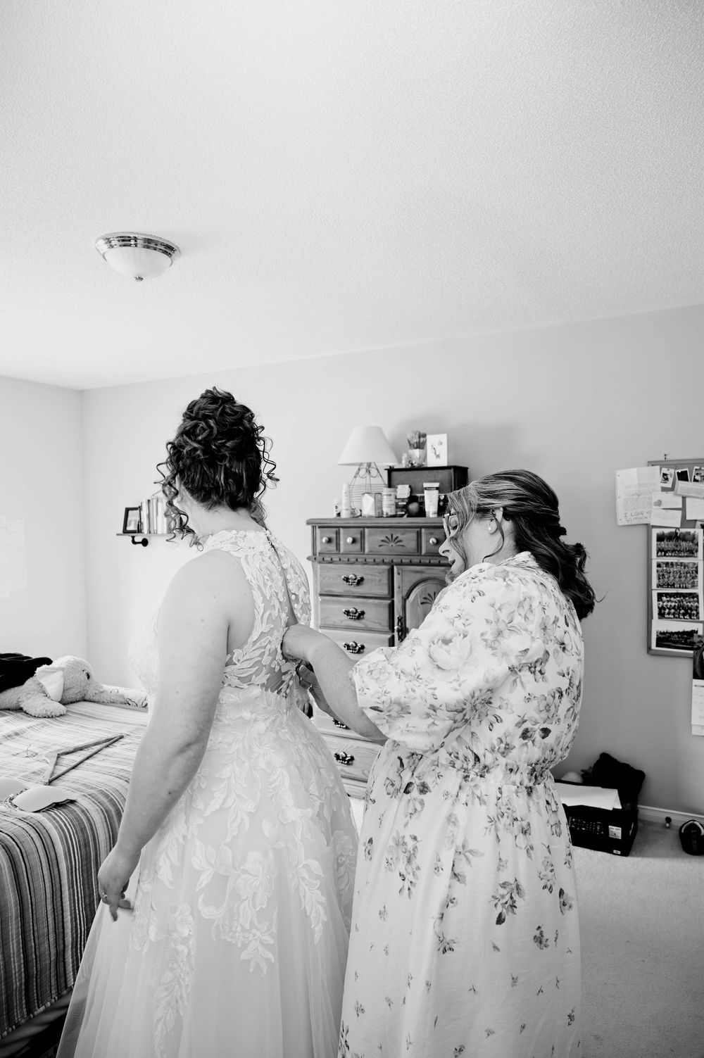Black and white photograph showing preparations for a wedding, with a bride in a lace dress being helped with final details.