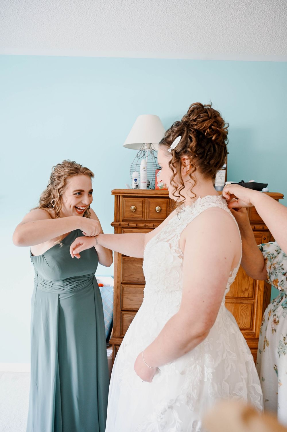 Two people share a special moment during wedding preparations in a room with sage green and white attire.