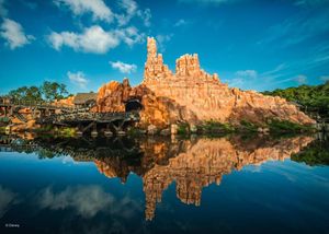 A panoramic view of red rock formations reflecting in calm water under vibrant blue skies at Big Thunder Mountain.