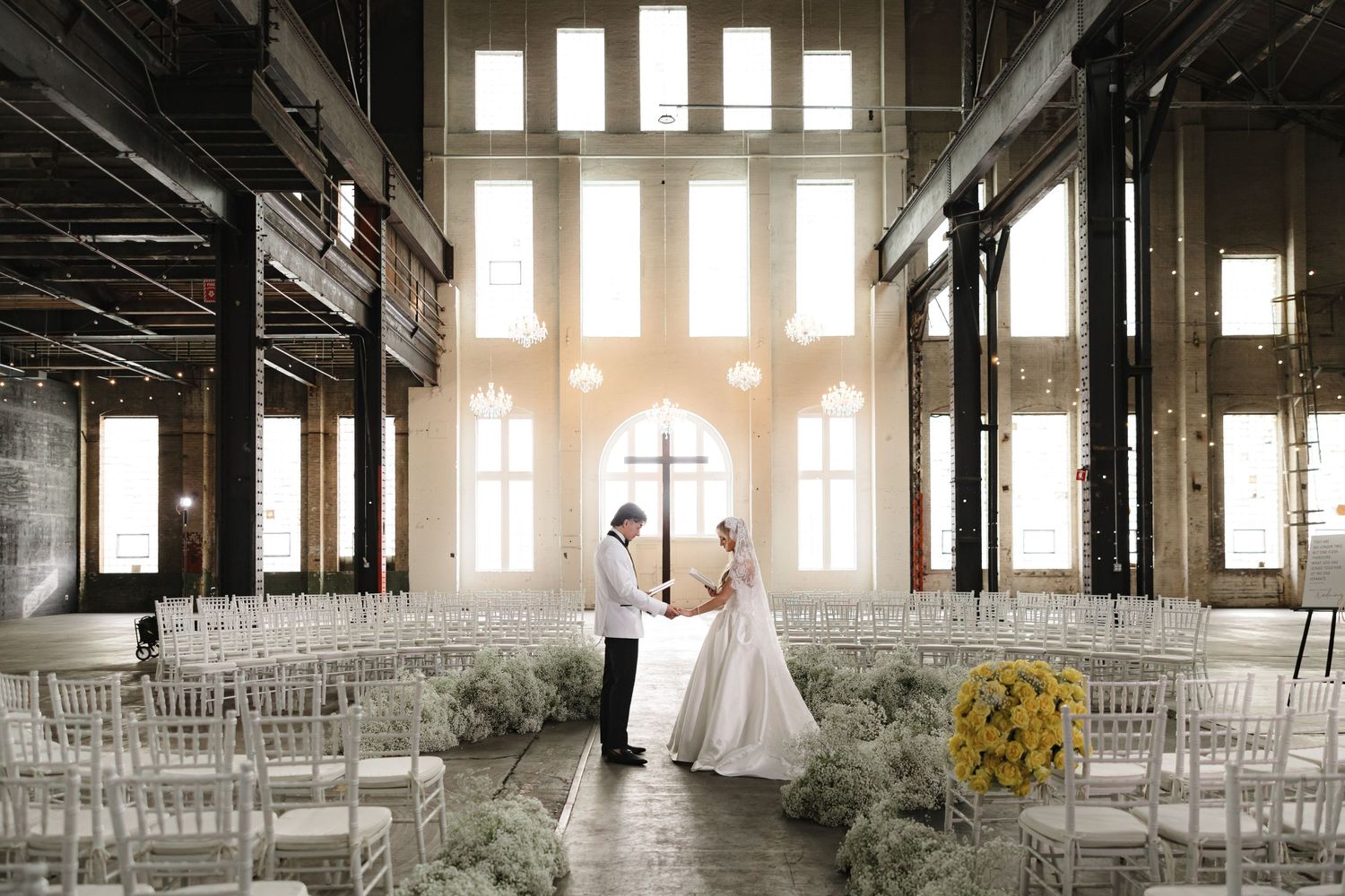 Minimalist wedding ceremony setting in industrial space with tall windows and ghost chairs.