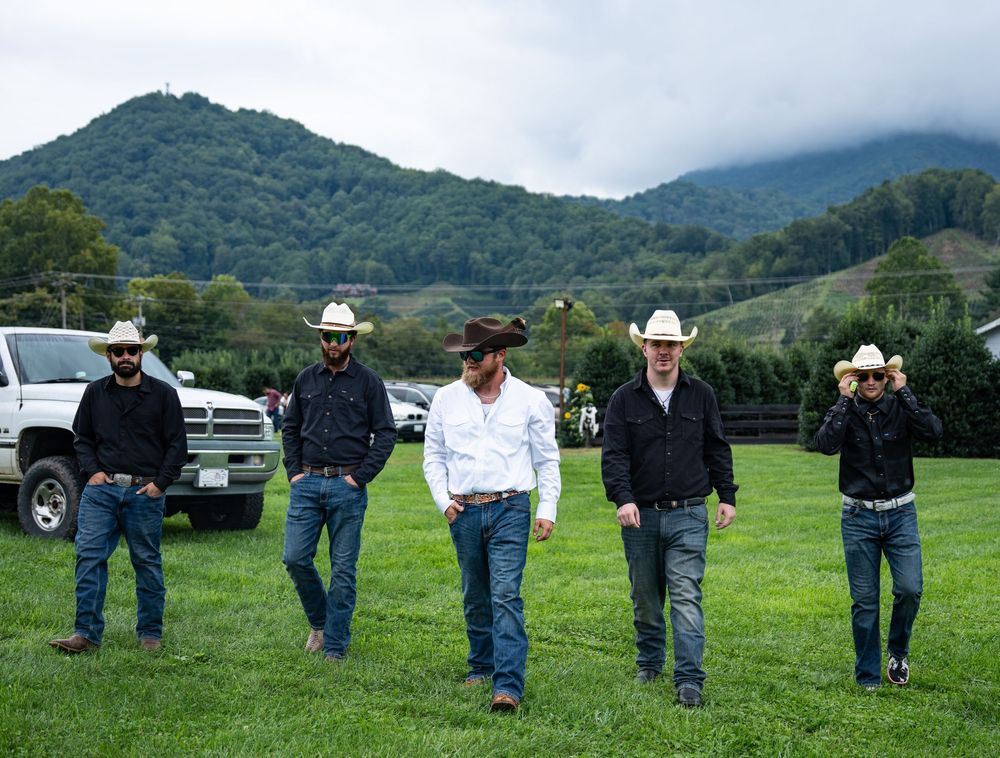 Five people in cowboy hats and western attire standing in a line on grass with mountains in the background.