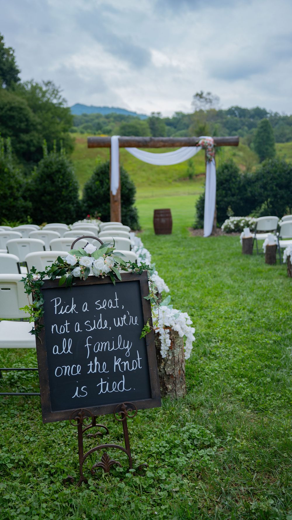 Rustic chalkboard wedding signs with white text displayed on grass at an outdoor ceremony venue with mountains in background.