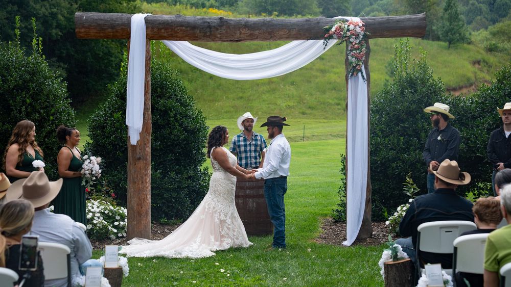 Outdoor wedding ceremony under a wooden arch draped with white fabric in a scenic countryside setting.