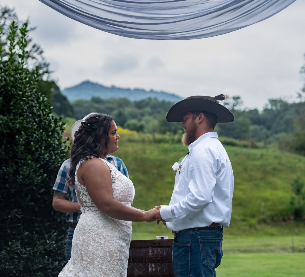 A couple in wedding attire exchanges vows during an outdoor ceremony with rolling hills in the background.