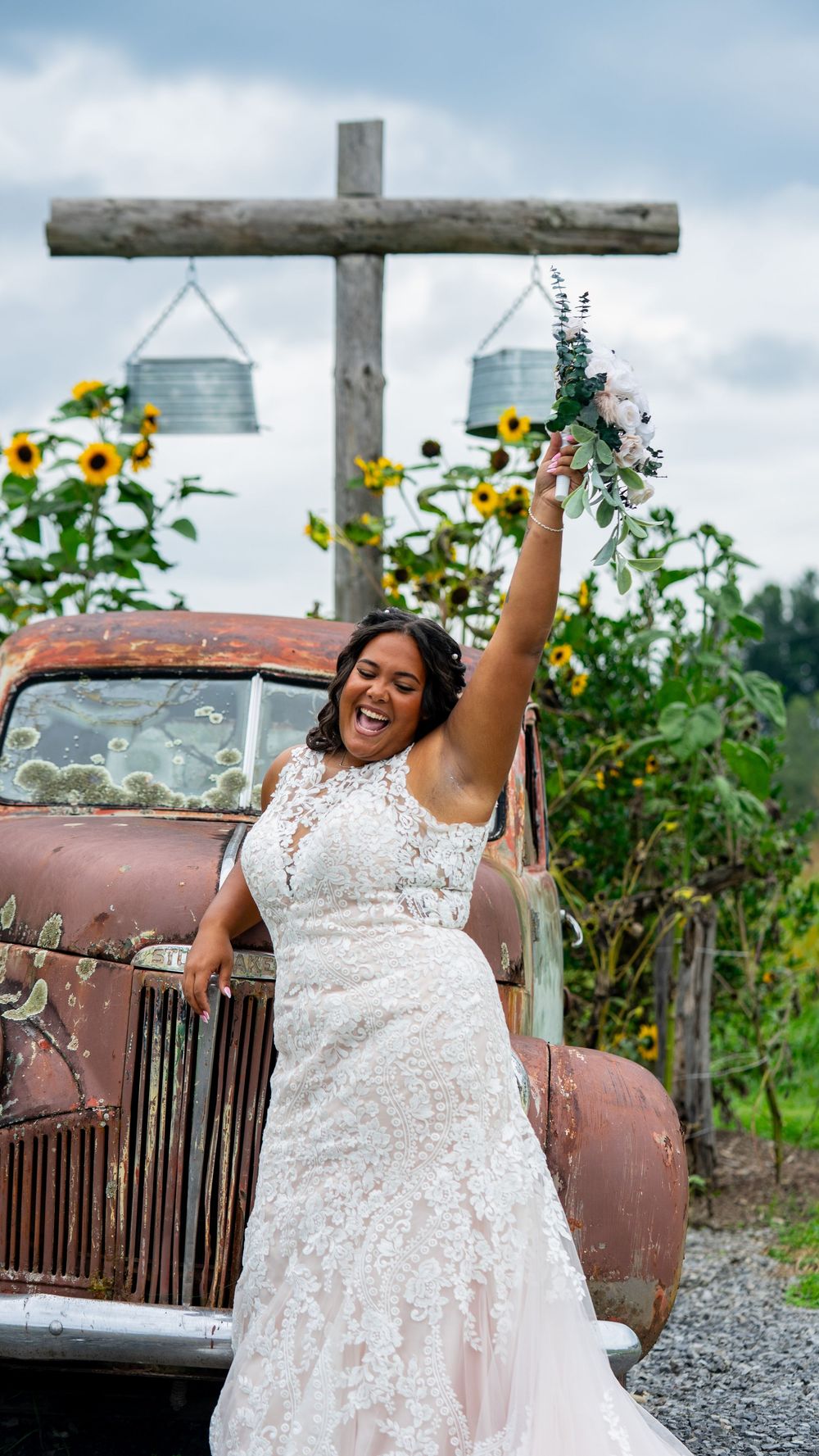 A joyful celebration in a white lace dress next to a vintage rustic truck with sunflowers and hanging decor in the background.