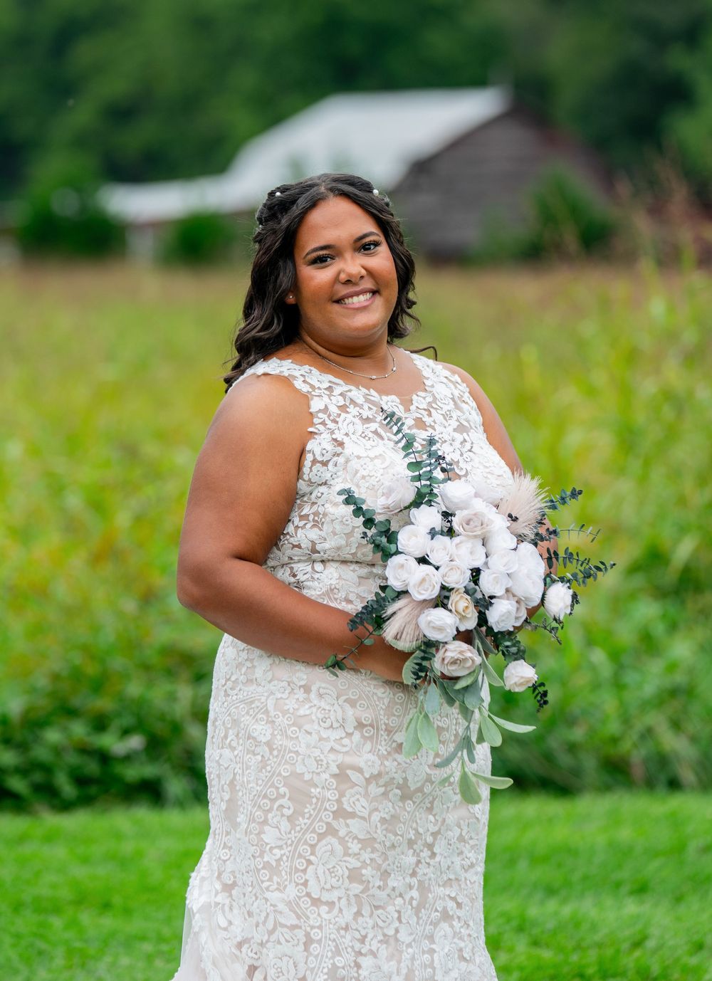A bride in a lace wedding dress poses with a beautiful white and pink floral bouquet in a green field.