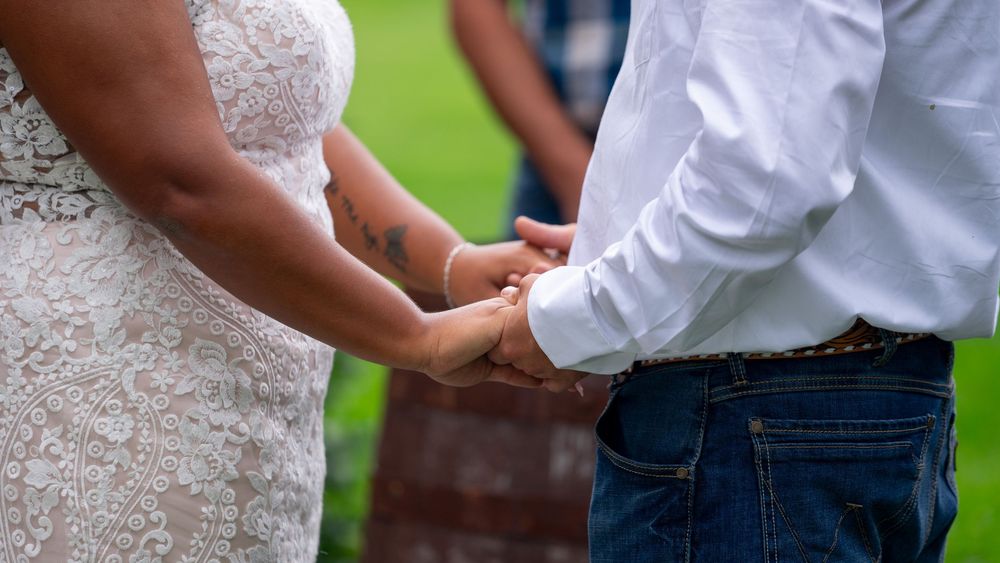 Close-up detail of wedding attire showing white dress and denim jeans with hands clasped together.