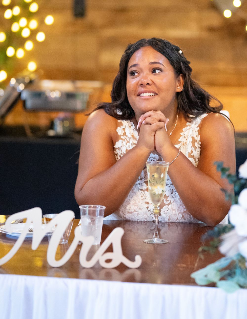 A person in a white lace dress smiles while sitting at a table with soft glowing lights in the background.