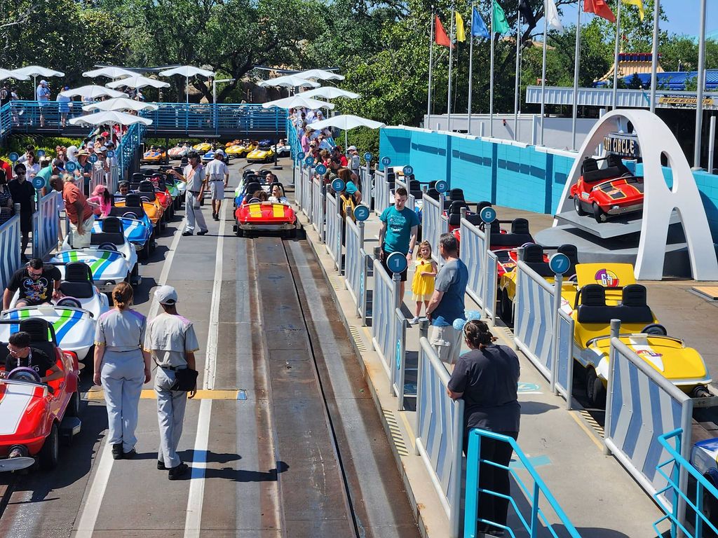 Theme park visitors board colorful Tomorrowland Speedway race cars under blue umbrellas at Disney park on a sunny day.