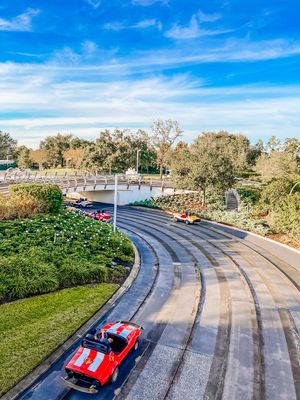Red and white race cars navigate a winding track with trees and blue skies at Walt Disney World's Tomorrowland Speedway.