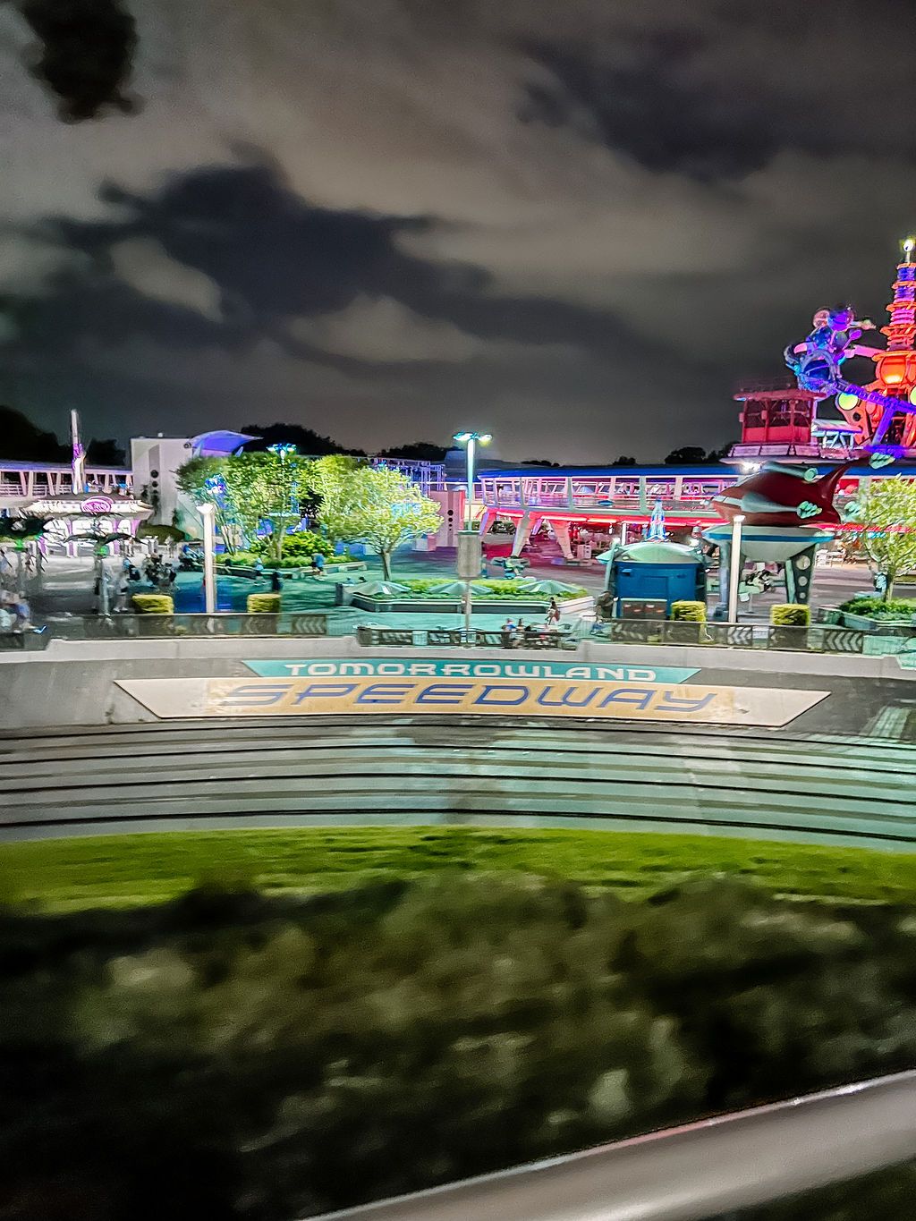 Vibrant neon-lit carnival rides and attractions illuminate an amusement park at night with a dramatic cloudy sky overhead.
