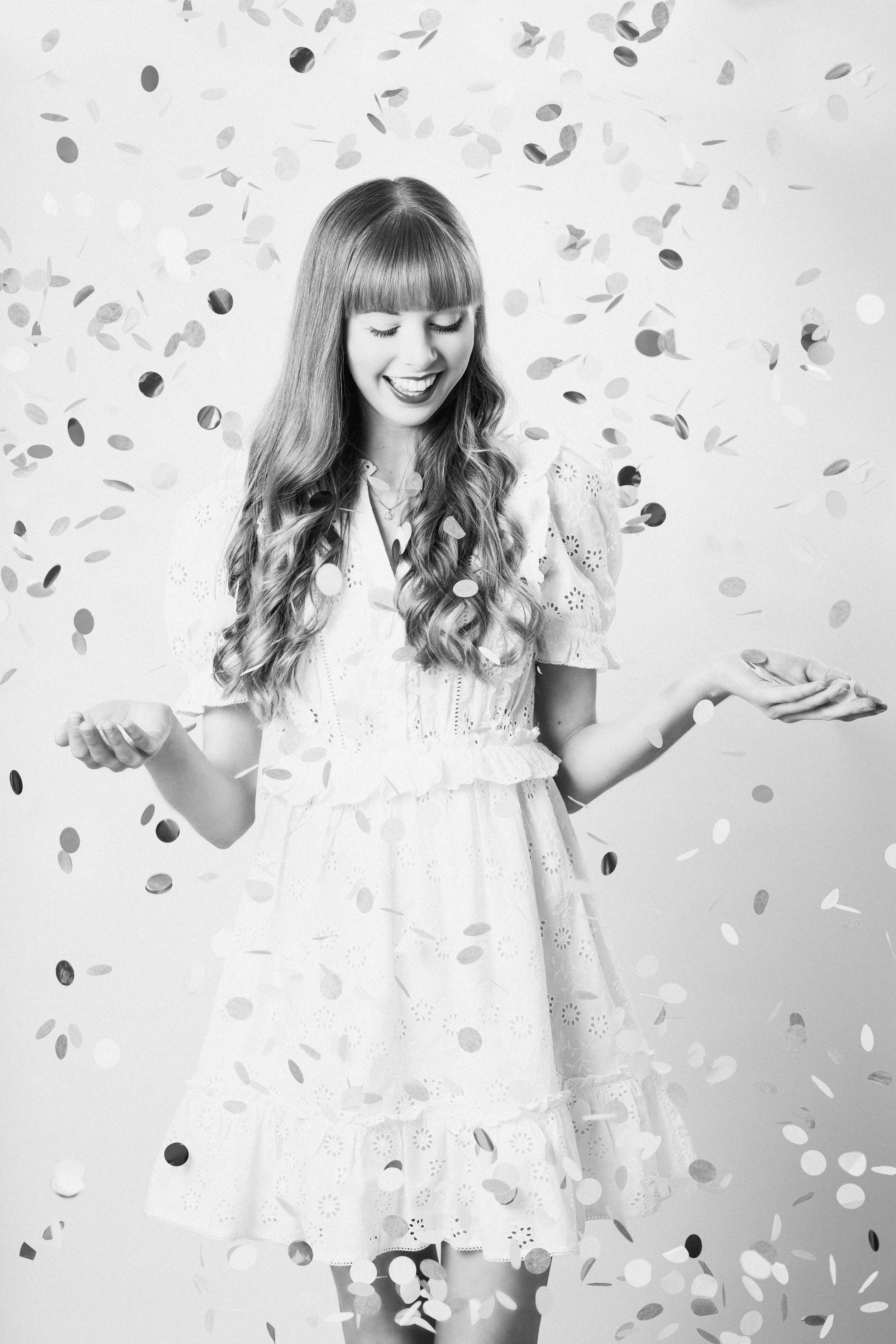A joyful figure in a white lace dress poses with hands raised while confetti falls against a black and white background. Wedding celebrant.