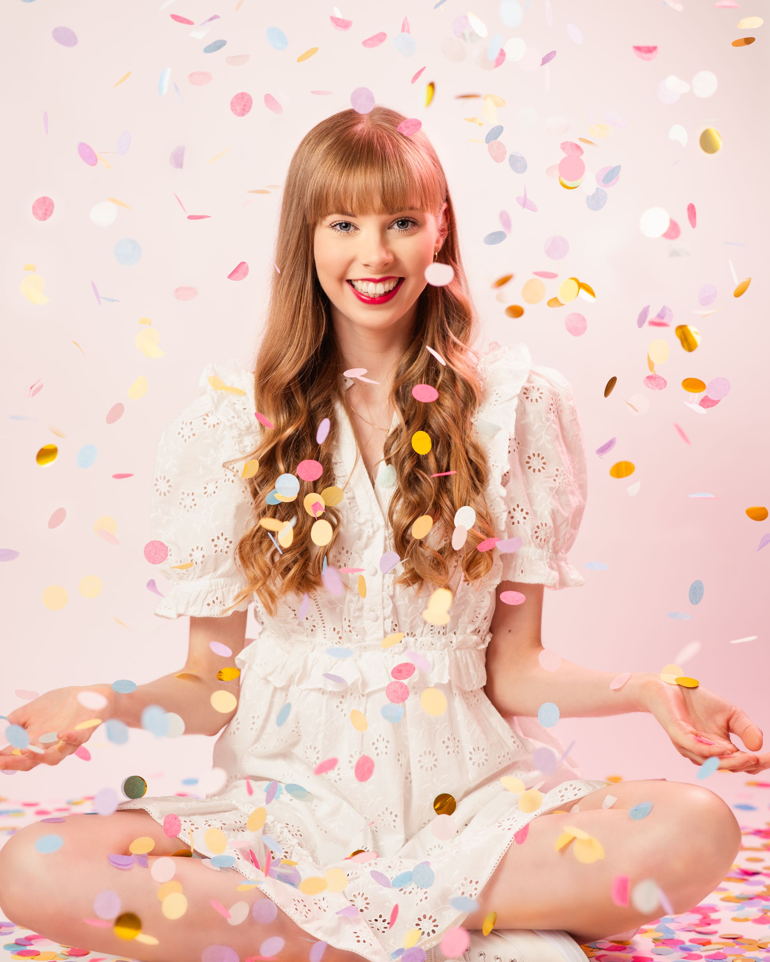 Colorful confetti rains down as someone in a white dress sits cross-legged on a pink background. Personal Branding Photography Surrey.