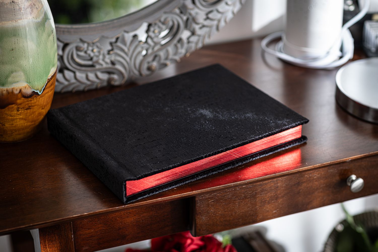 A black suede album with red gilded pages sits on a dark wooden table with decorative elements nearby.