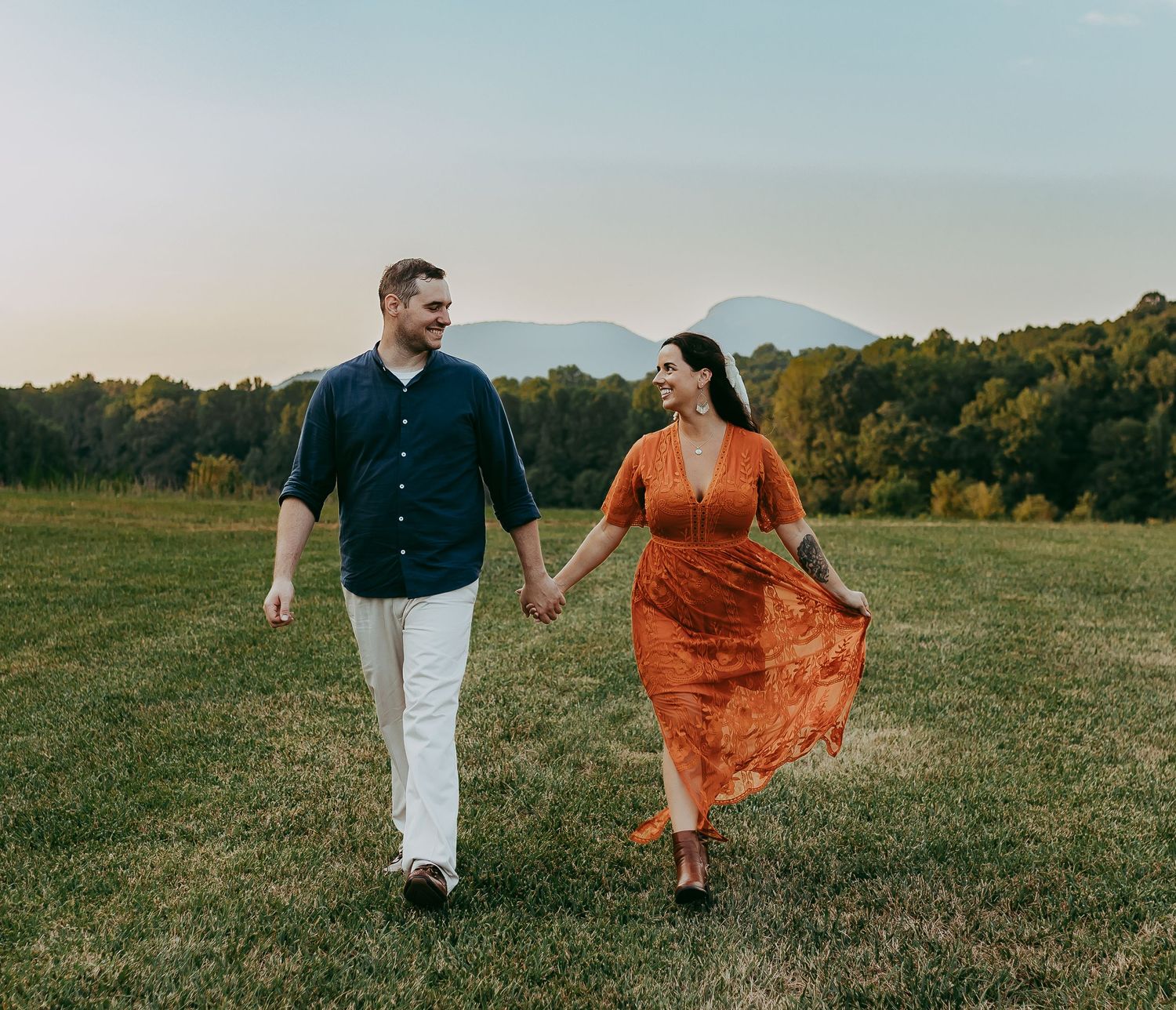 A couple walks hand in hand through a grassy field at sunset with the woman wearing a flowing rust-colored dress.