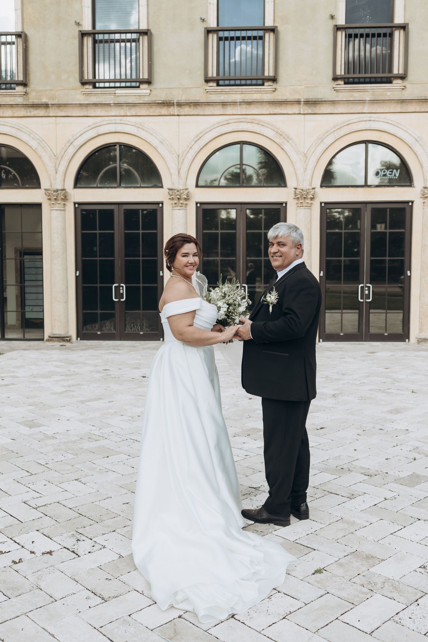 Bride in flowing white gown poses with father outside elegant arched building.