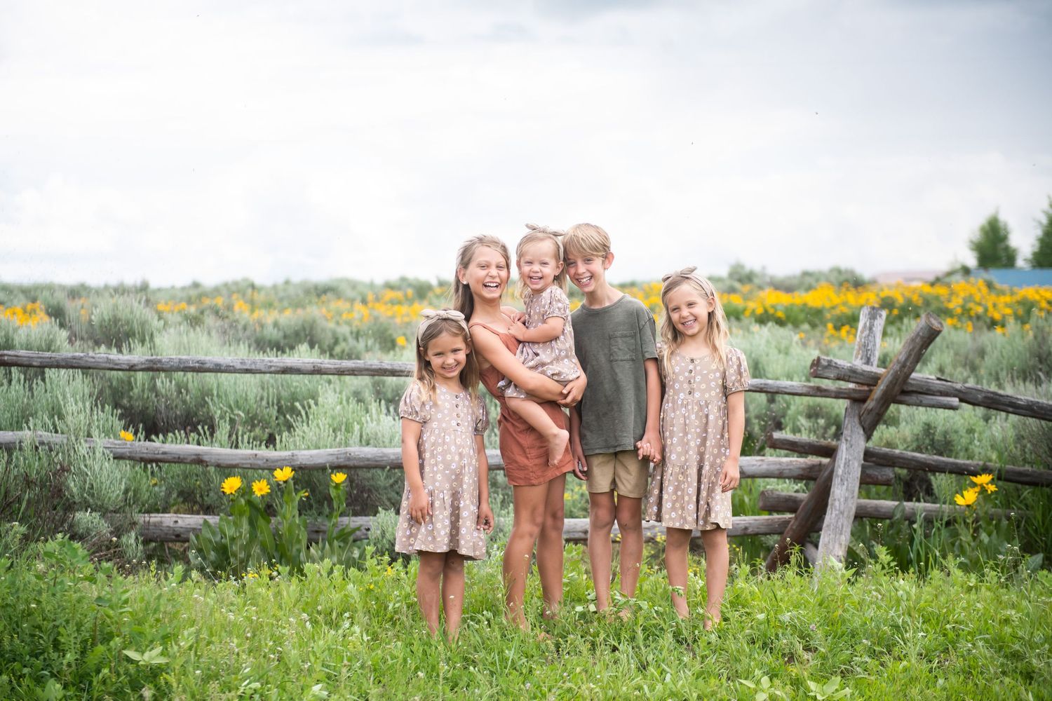 Children standing together by wooden fence with yellow wildflowers blooming in background.
