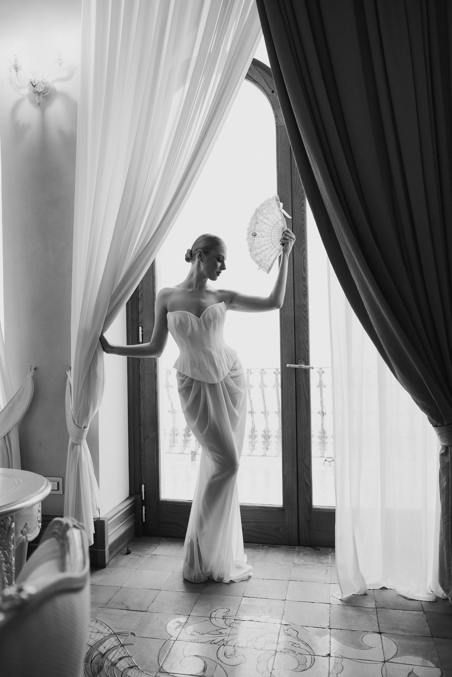 Black and white photo of elegant bride in strapless gown holding fan by ornate window curtains.