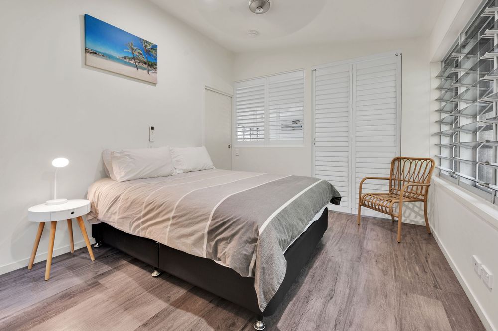 Minimalist bedroom with white walls, hardwood flooring, and natural light streaming through shuttered windows.