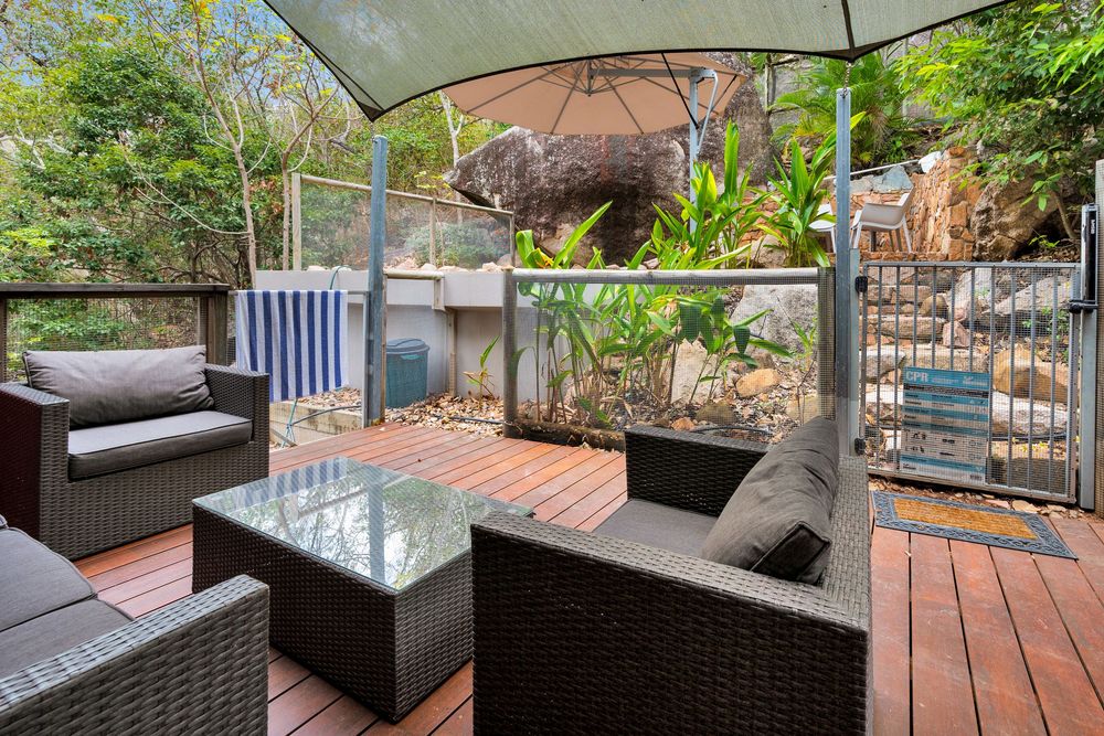Outdoor patio deck with rattan furniture and glass railings surrounded by lush greenery and trees.