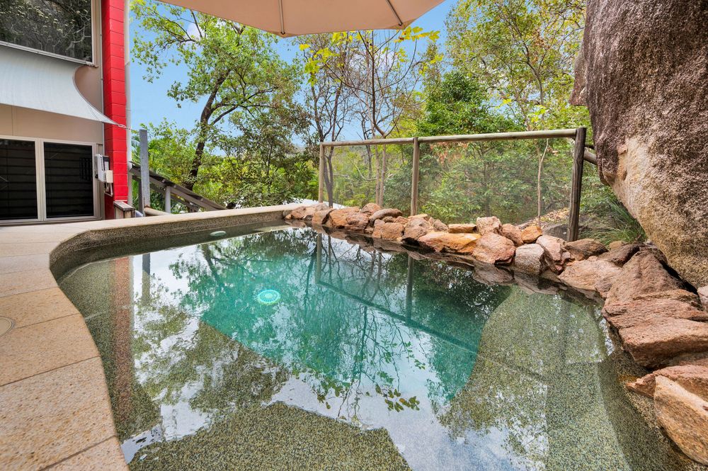 Tranquil swimming hole surrounded by rocky cliffs and lush trees reflects clear blue water on a sunny day.