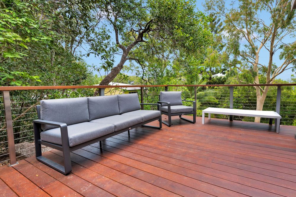 Panoramic view of a modern wooden deck with grey outdoor furniture and metal railings surrounded by lush green trees.