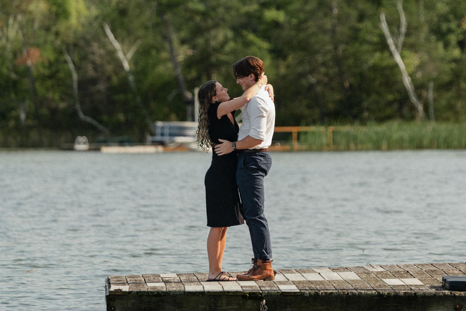 A romantic couple stands embracing on a wooden dock by a tranquil lake surrounded by trees.