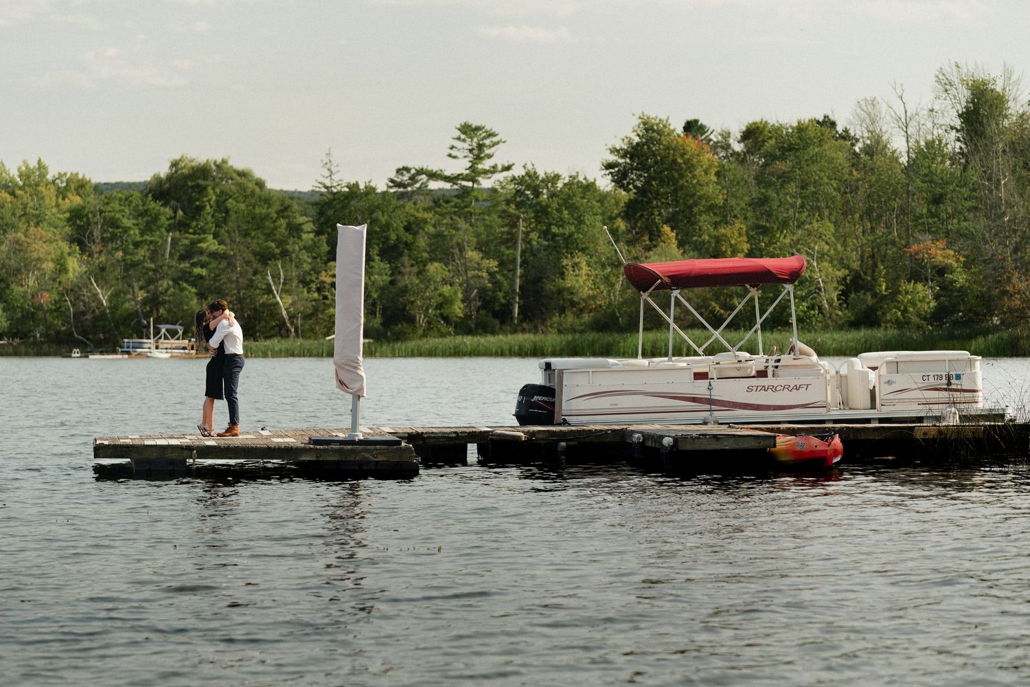 Two people stand on a floating dock with a pontoon boat moored nearby on a serene lake.