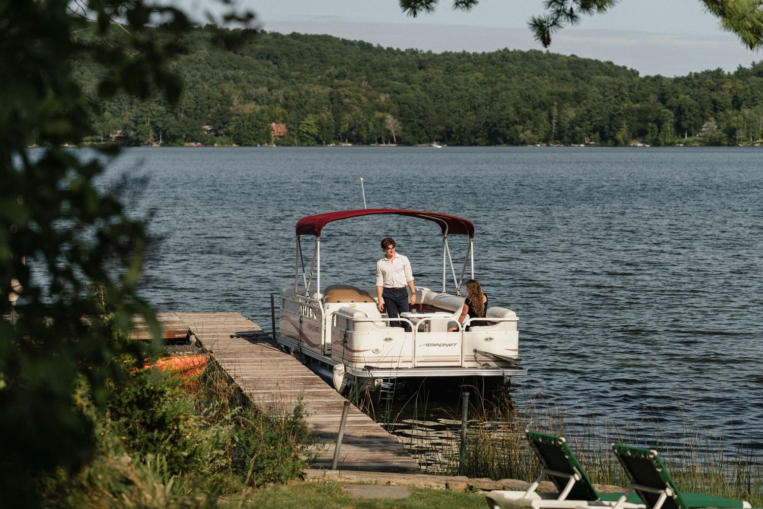 A moored pontoon boat with red canopy sits at a lakeside dock surrounded by green hills.