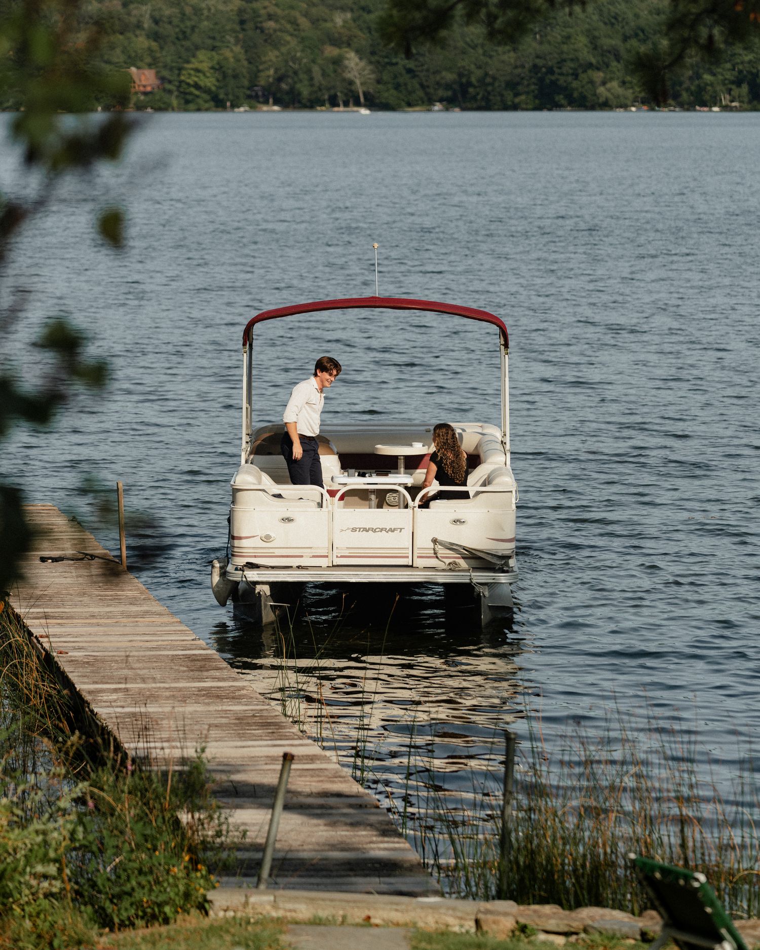 A white pontoon boat with red canopy is tied to a wooden dock on a scenic lake.