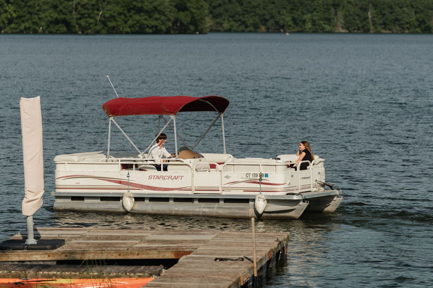 A white pontoon boat with red canopy cruises near a wooden dock on a summer lake.