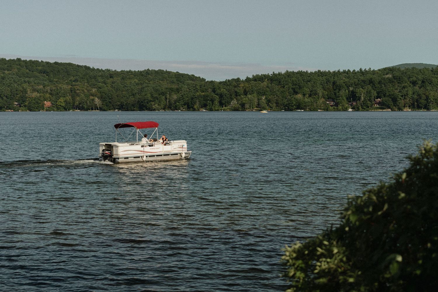 A pontoon boat with red sunshade cruises across a scenic lake surrounded by wooded hills.