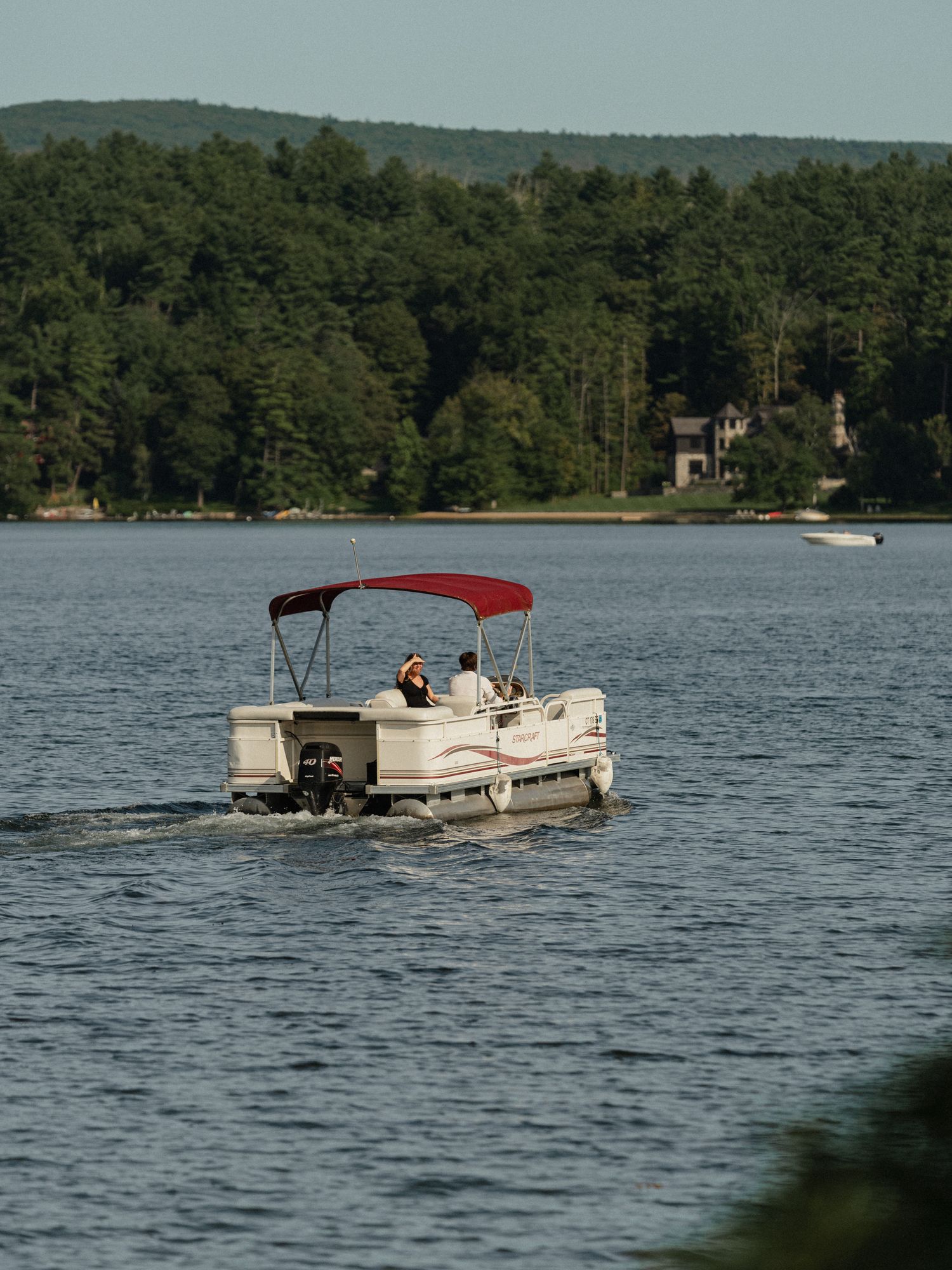A white pontoon boat glides across calm lake waters with forested shores in the background.