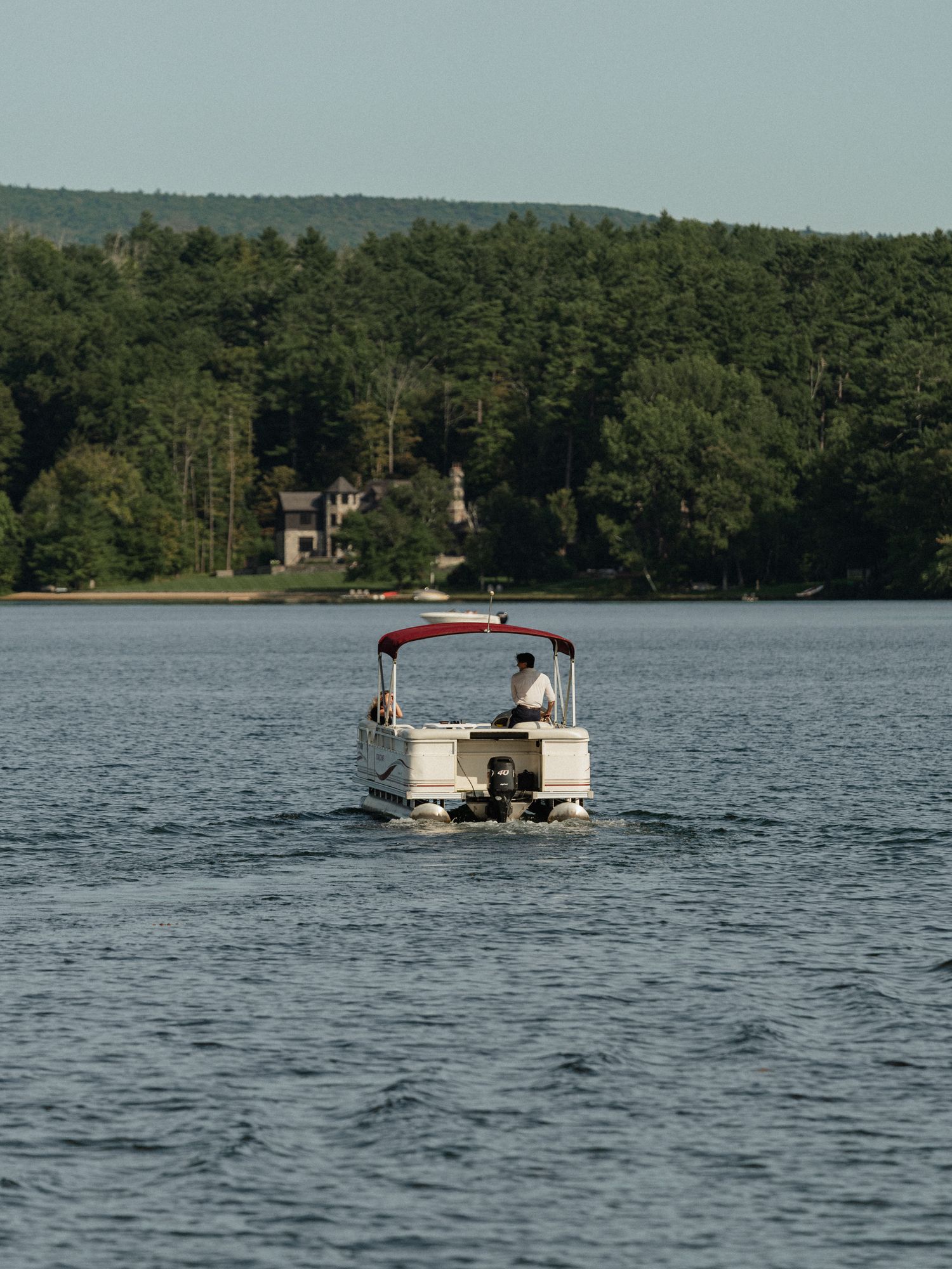 A pontoon boat navigates across a serene lake with lakefront homes visible through the trees.
