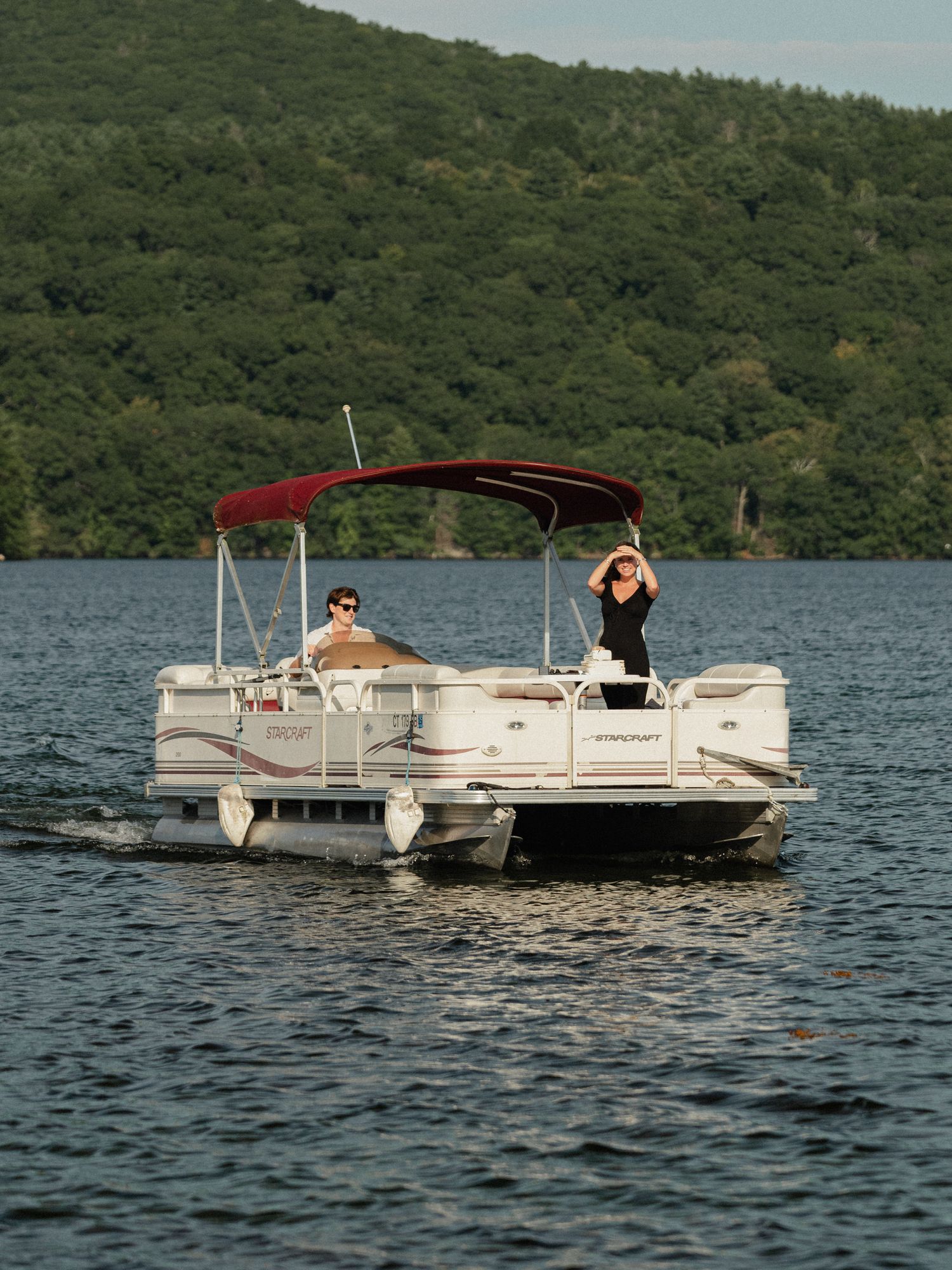 A pontoon boat floats on a lake surrounded by green forested hills on a sunny day.